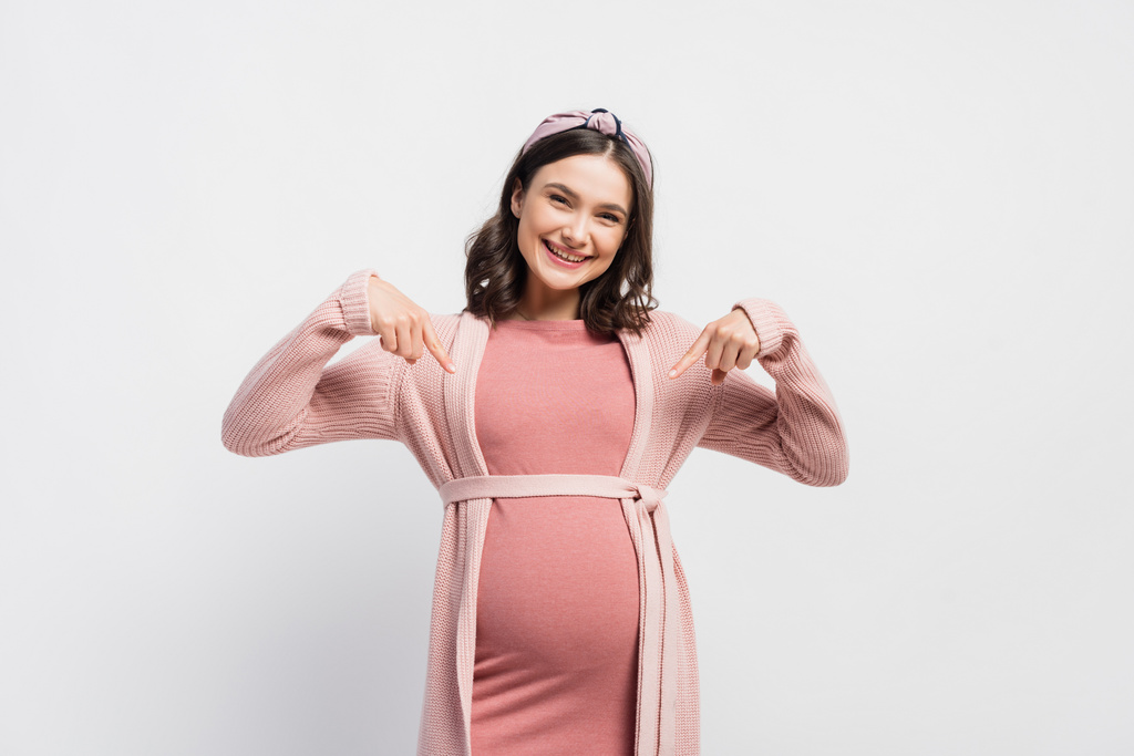 mujer alegre y embarazada señalando con los dedos el vientre sobre blanco - Foto, Imagen