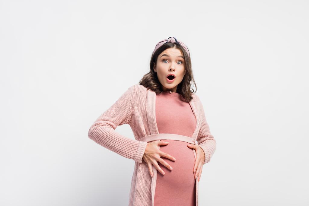 geschockte und schwangere Frau blickt in Kamera und berührt Bauch auf Weiß - Foto, Bild