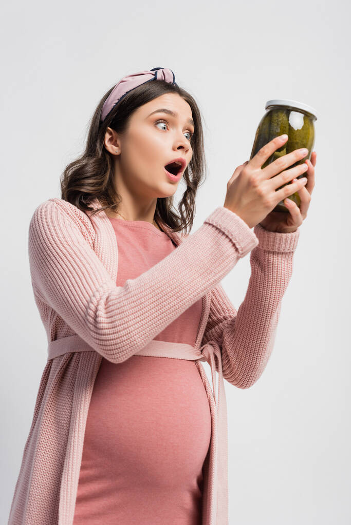 mujer sorprendida y embarazada sosteniendo frasco con pepinos en escabeche aislados en blanco - Foto, Imagen
