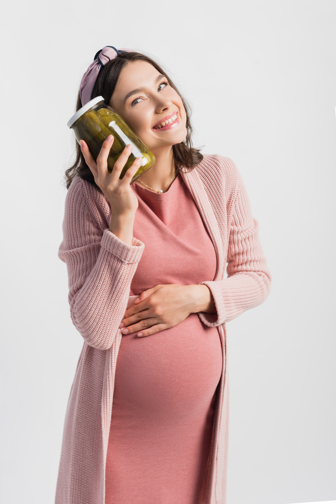 mujer satisfecha y embarazada sosteniendo frasco con pepinos en escabeche y mirando hacia otro lado aislado en blanco - Foto, Imagen