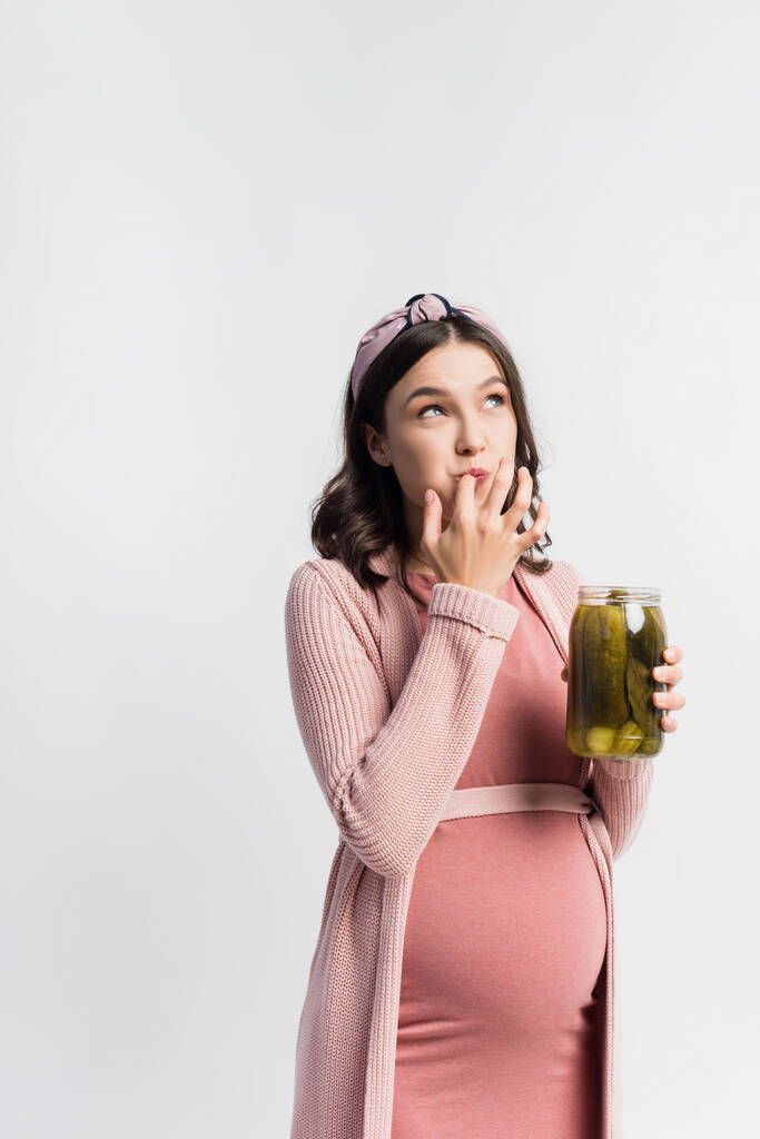 mujer embarazada sosteniendo frasco con pepinos en escabeche mientras lame el dedo aislado en blanco - Foto, imagen