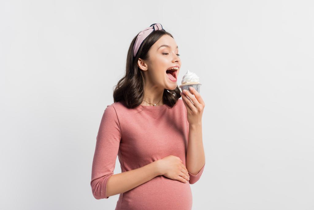 gioiosa donna incinta con la bocca aperta che tiene cupcake isolato su bianco - Foto, immagini