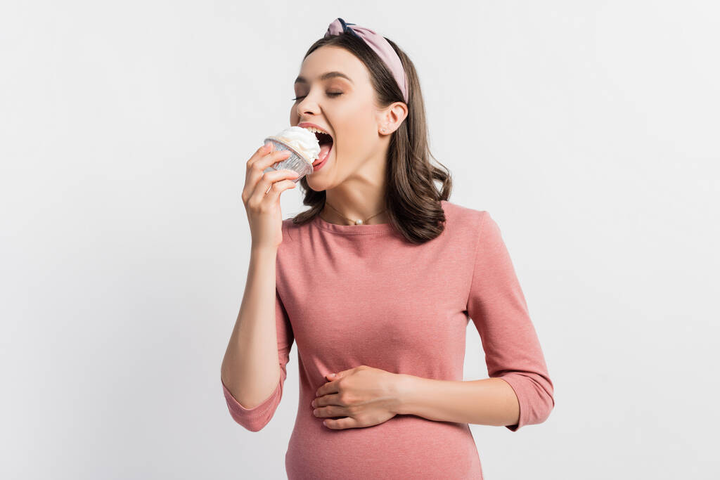vrolijke zwangere vrouw met gesloten ogen eten cupcake geïsoleerd op wit - Foto, afbeelding