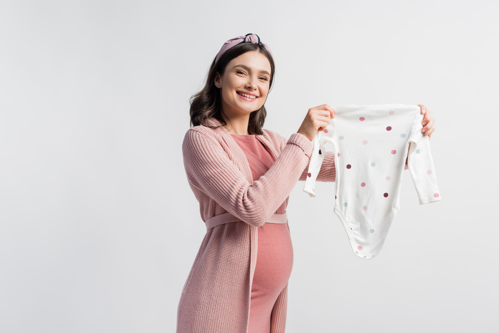mujer embarazada complacida en diadema sosteniendo bebé mameluco aislado en blanco  - Foto, imagen