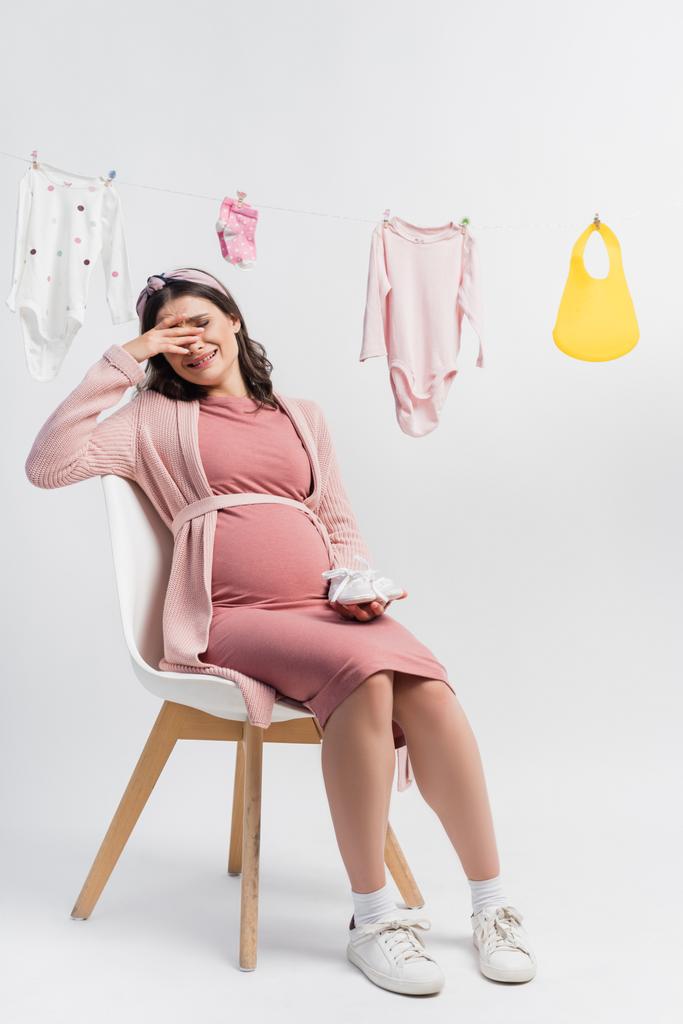 donna incinta seduta sulla sedia e piangendo vicino a vestiti per bambini su bianco - Foto, immagini
