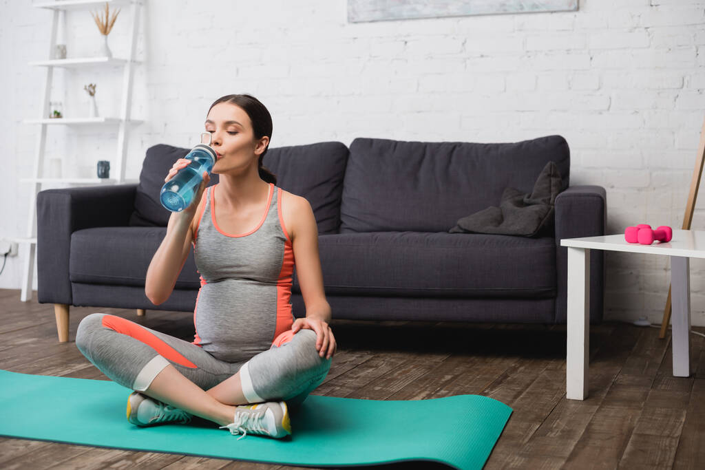 ciężarna kobieta siedzi na macie fitness podczas picia wody z butelek sportowych - Zdjęcie, obraz