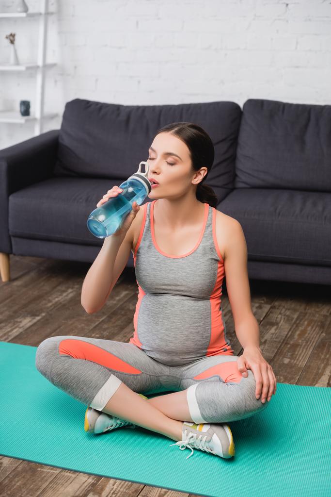妊娠中の女性は水を飲みながらスポーツボトルを持ってフィットネスマットに座って - 写真・画像