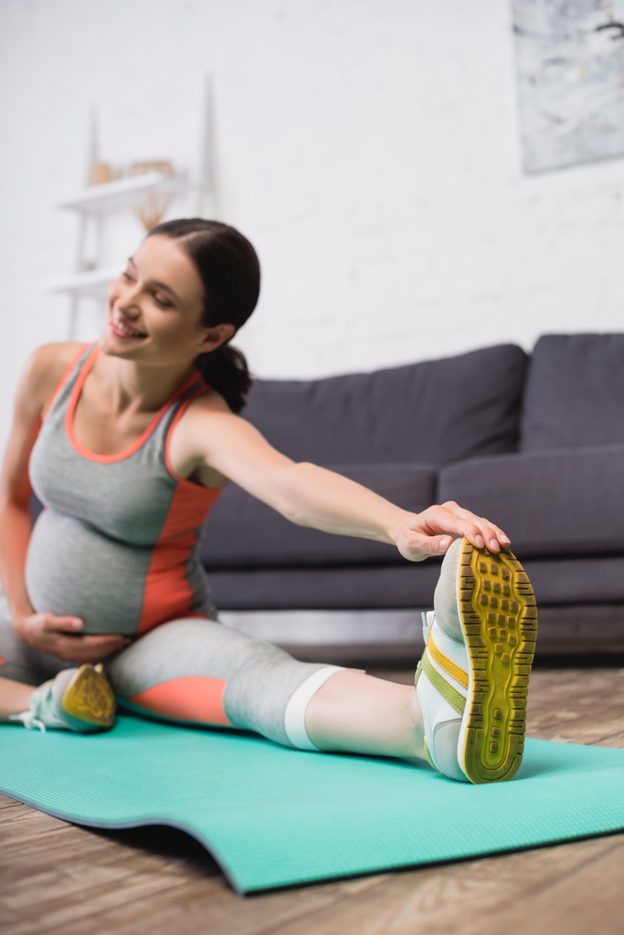 Spor giyim kuşamında spor kıyafetli neşeli hamile kadının spor paspasına esnediği seçici odak noktası - Fotoğraf, Görsel