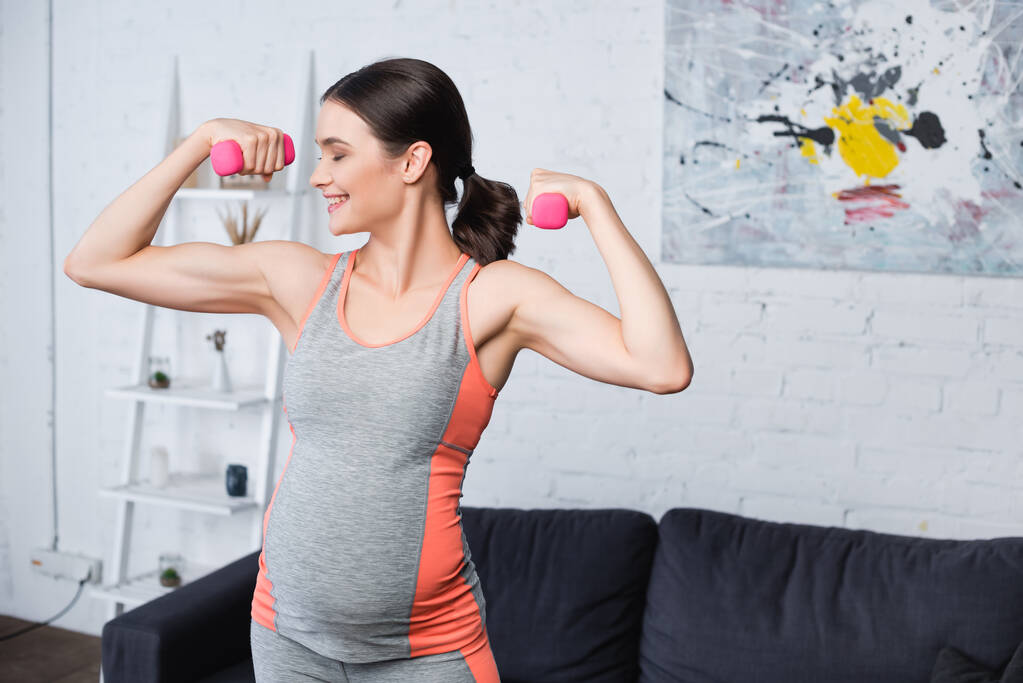 freudige Schwangere trainiert zu Hause mit pinkfarbenen Hanteln - Foto, Bild
