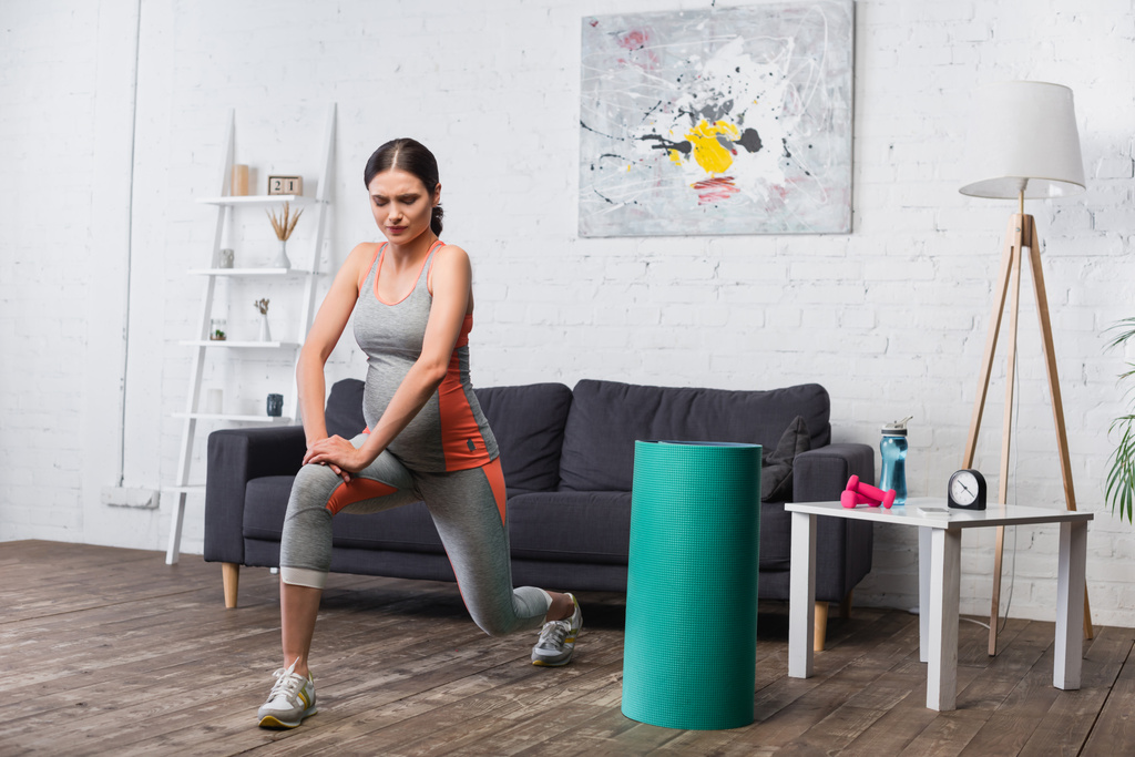 bruneta a těhotná žena cvičení v blízkosti sportovního vybavení v obývacím pokoji - Fotografie, Obrázek