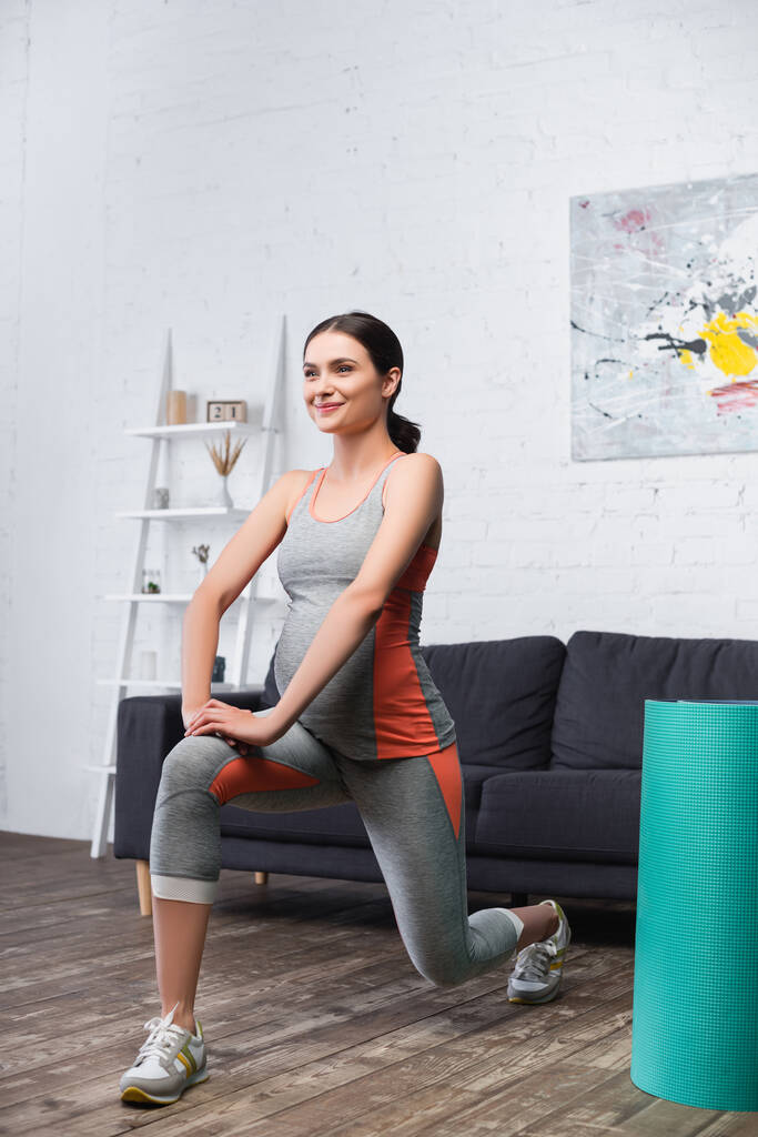 femme joyeuse et enceinte faisant de l'exercice près tapis de fitness dans le salon - Photo, image