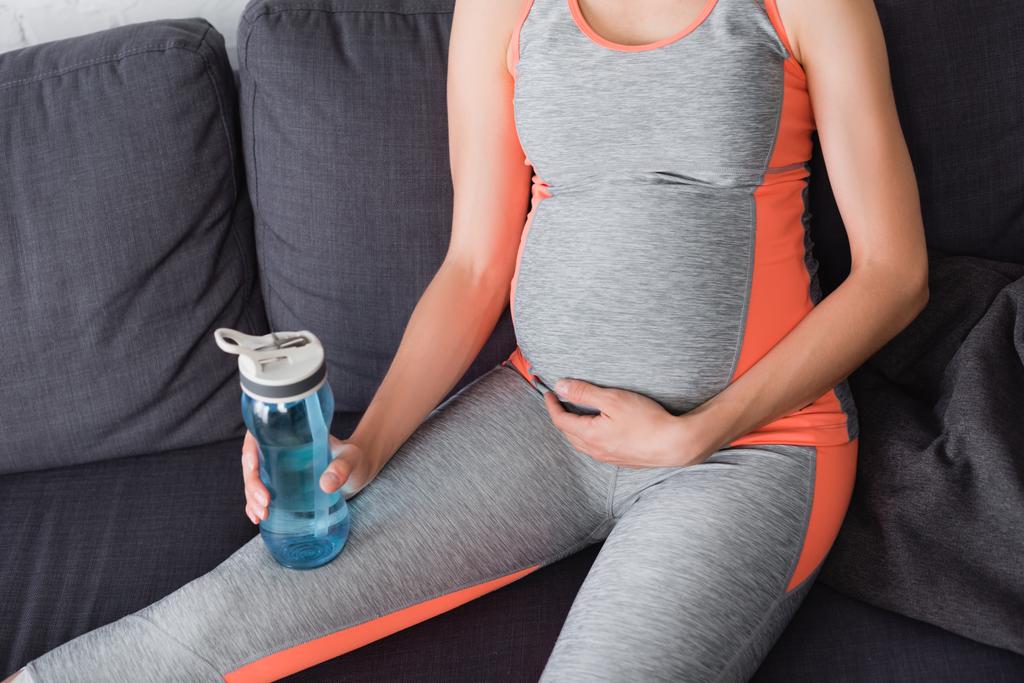 rajattu näkymä raskaana istuva nainen sohvalla ja tilalla urheilu pullo vedellä - Valokuva, kuva