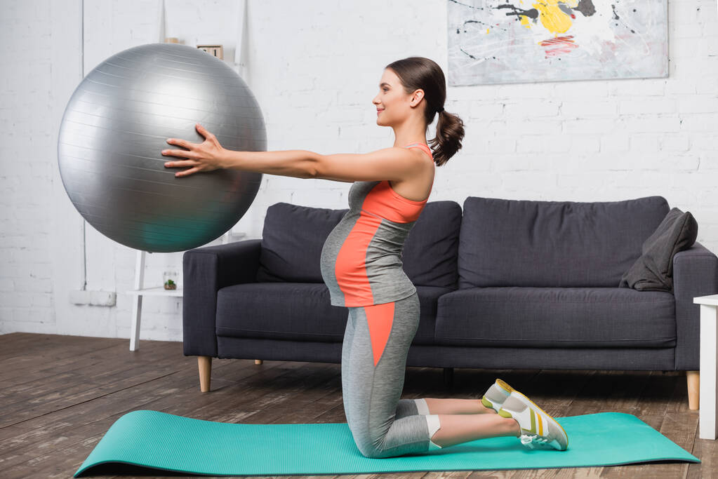 visão lateral da mulher grávida trabalhando com bola de fitness na sala de estar  - Foto, Imagem