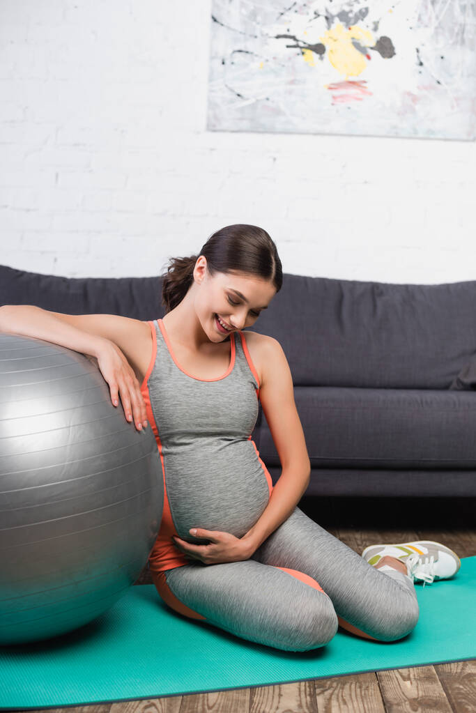 Freudige und schwangere Frau berührt Bauch, während sie in der Nähe von Fitnessball im Wohnzimmer sitzt  - Foto, Bild