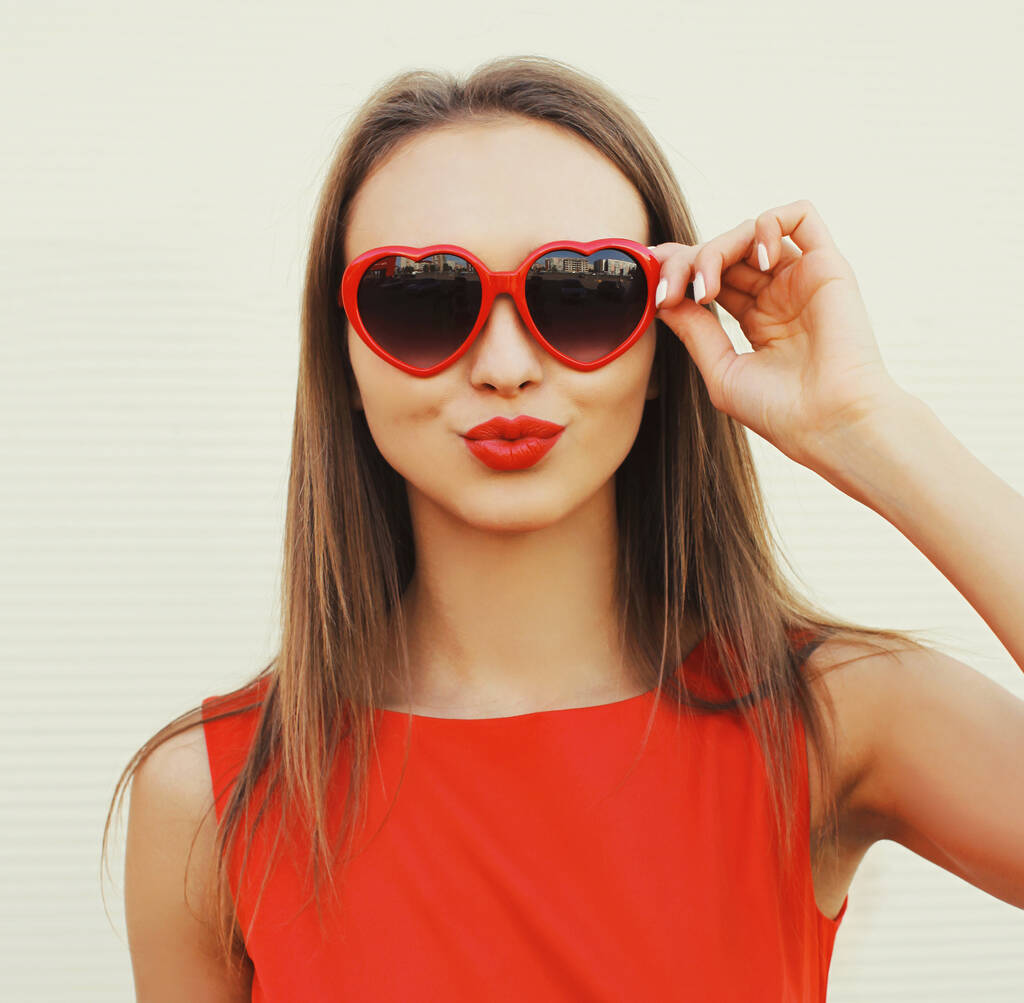 Retrato de cerca de una atractiva joven que sopla labios rojos enviando un beso de aire dulce con gafas de sol en forma de corazón sobre fondo blanco - Foto, imagen