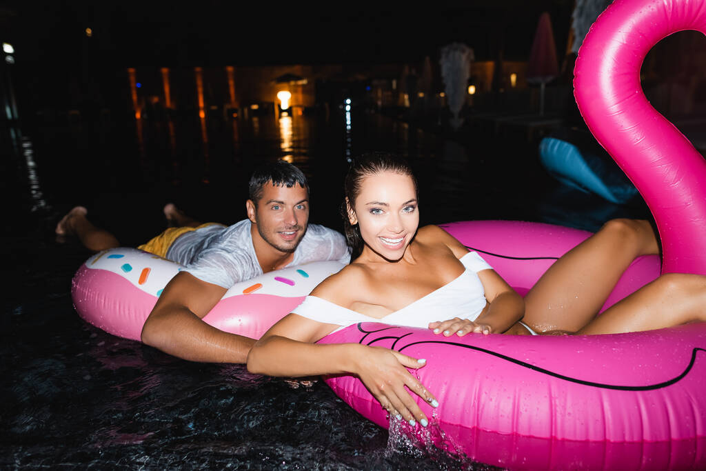 Selective focus of young couple looking at camera while swimming on rings in pool at night  - Photo, Image
