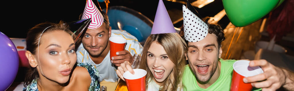 Panoramic orientation of excited friends in party caps holding disposable cups near balloons at night  - Photo, Image
