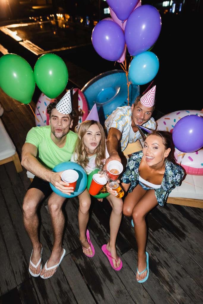 Νεαροί φίλοι κοιτάζουν την κάμερα ενώ κάνουν πρόποση με μπύρα κοντά σε μπαλόνια και πισίνα το βράδυ  - Φωτογραφία, εικόνα