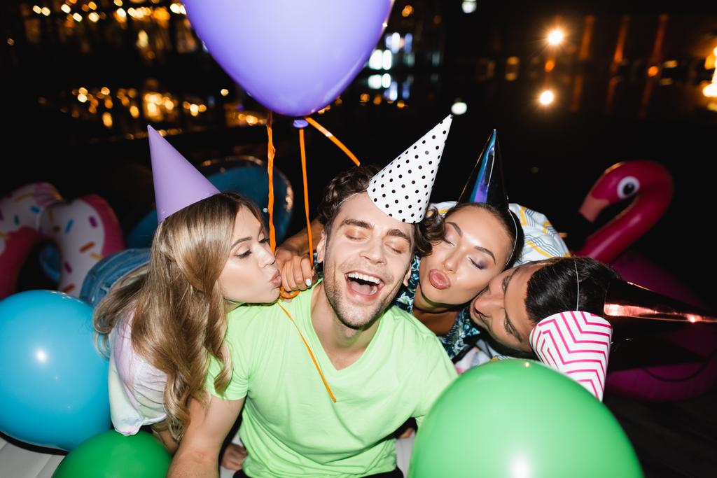 Selektywne skupienie przyjaciół w czapki party całuje człowieka z balonami na zewnątrz w nocy  - Zdjęcie, obraz