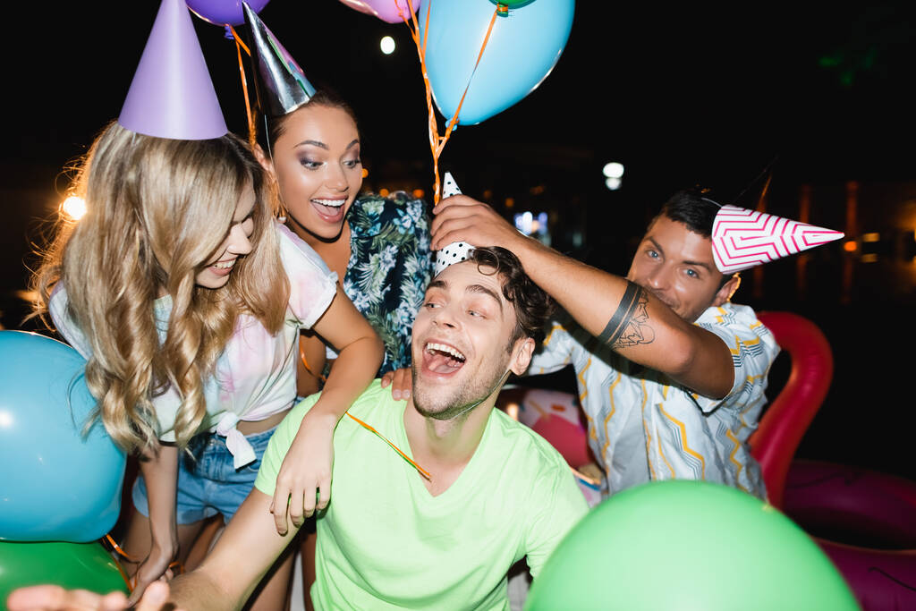 Enfoque selectivo del hombre que usa gorra de fiesta en la cabeza de un amigo cerca de las mujeres y globos por la noche  - Foto, imagen