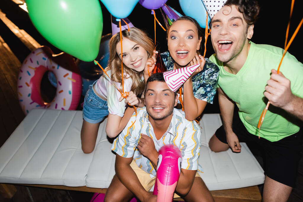 Mladí přátelé ve slavnostních čepicích drží balónky v blízkosti plaveckého kroužku v noci  - Fotografie, Obrázek