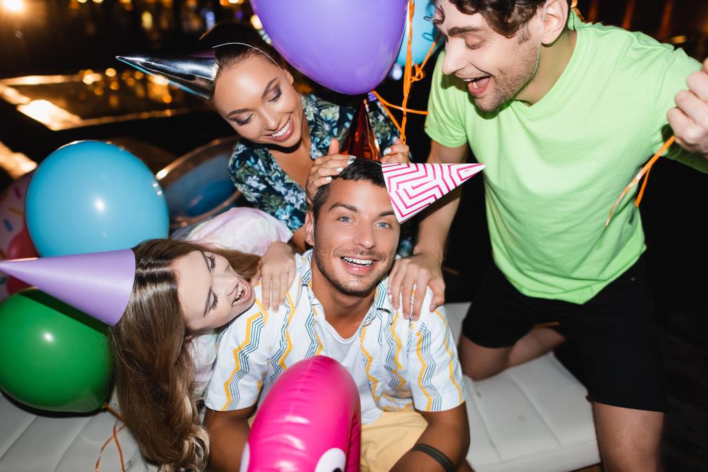 Selectieve focus van vrienden knuffelen jongeman in party cap in de buurt van ballonnen en zwembad 's nachts  - Foto, afbeelding