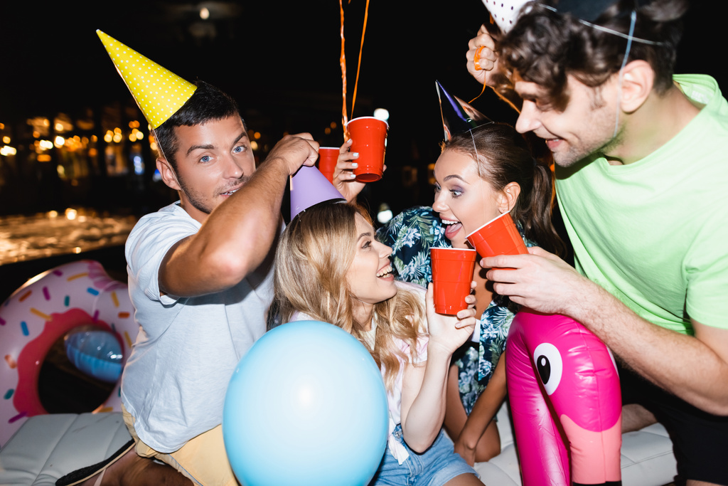 Enfoque selectivo de amigos brindando con vasos desechables cerca de globos durante la fiesta por la noche  - Foto, Imagen