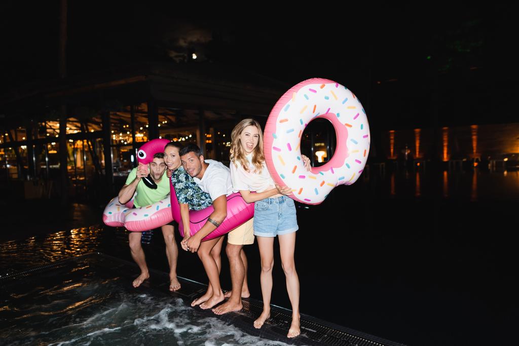 Захоплені молоді друзі з плавальними кільцями дивляться на камеру під час вечірки біля басейну вночі
  - Фото, зображення