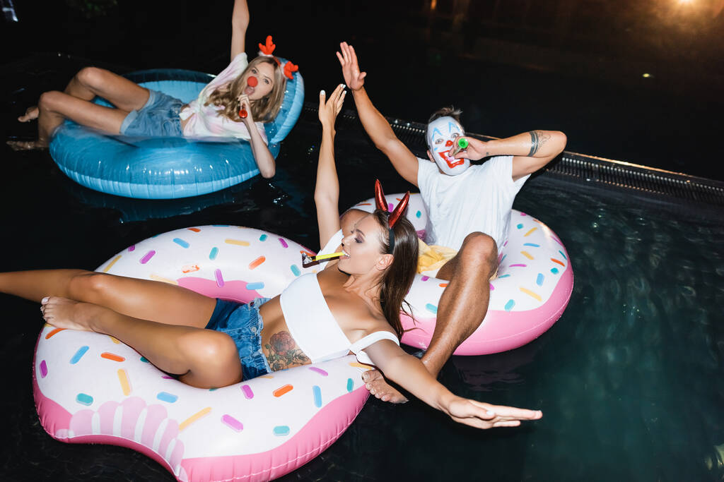 Вибірковий фокус друзів у масці та вечірніх головних уборах з дискетами, які плавають на кільцях у басейні вночі
  - Фото, зображення