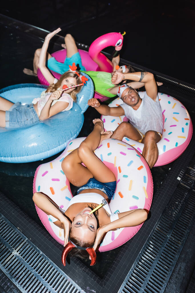 Wysoki kąt widzenia kobiety w party opaska z blow tickler pływanie na ringu w pobliżu przyjaciół w basenie w nocy  - Zdjęcie, obraz