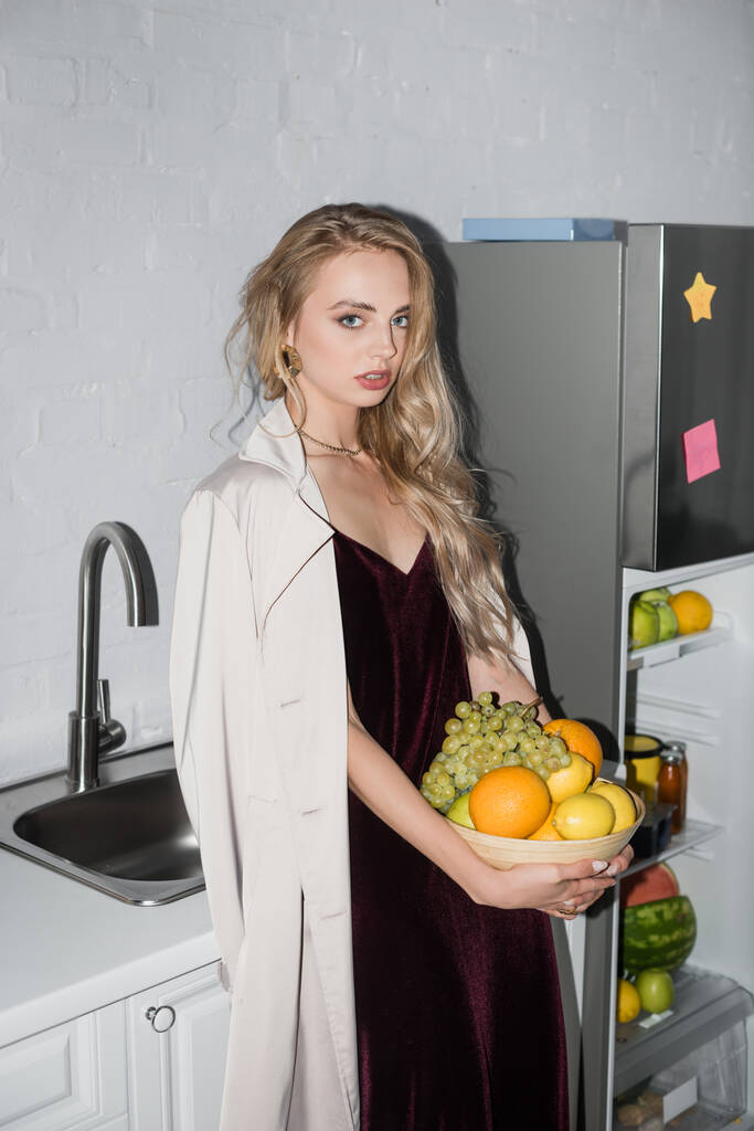 young blonde woman in trench coat and velour dress looking at camera while holding  bowl with fresh fruits - Photo, Image