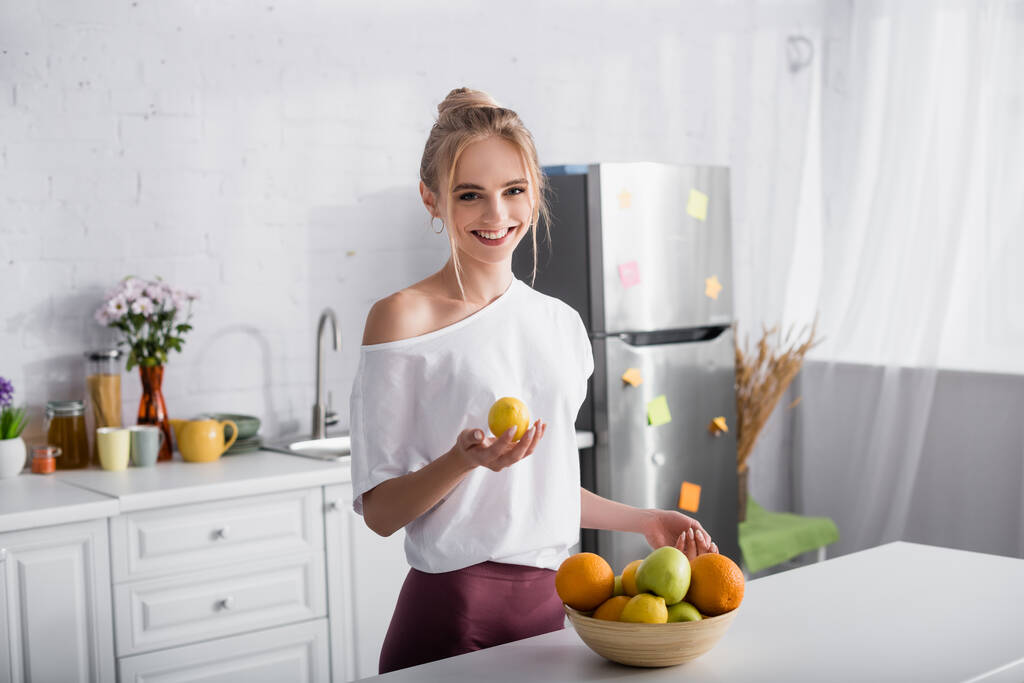 χαμογελαστή ξανθιά γυναίκα κρατώντας λεμόνι, ενώ στέκεται κοντά μπολ με φρέσκα φρούτα - Φωτογραφία, εικόνα
