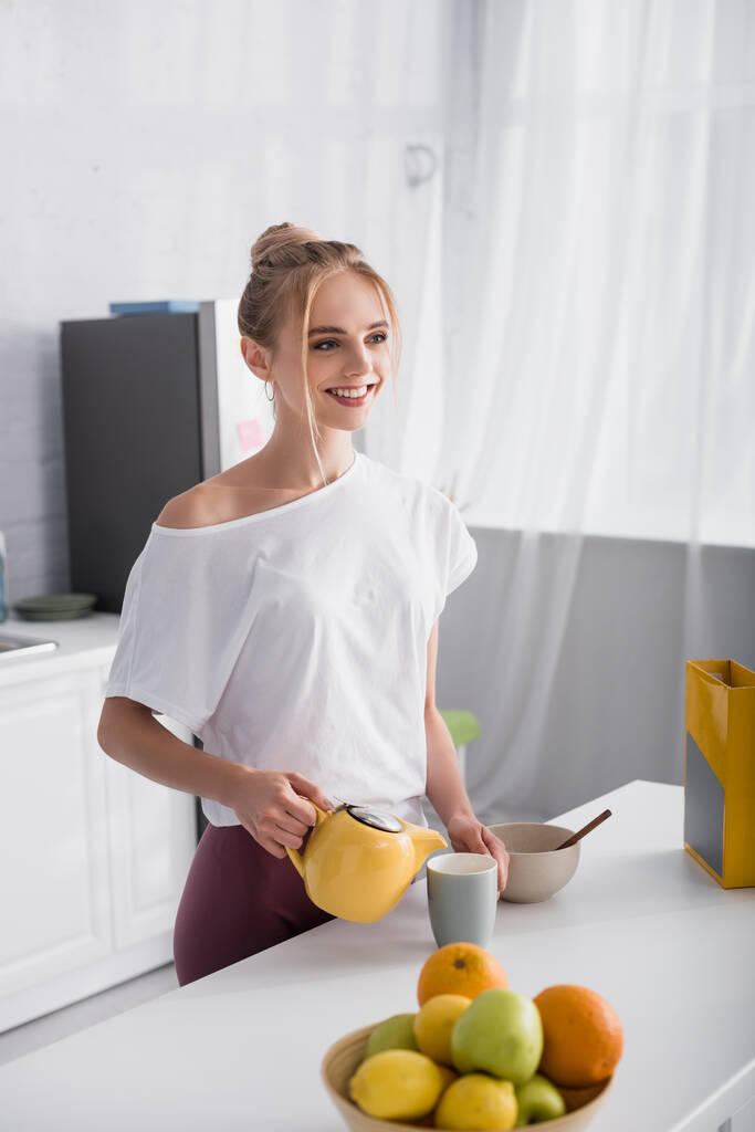 šťastná žena v bílém tričku dívá pryč při nalévání čaje do šálku - Fotografie, Obrázek