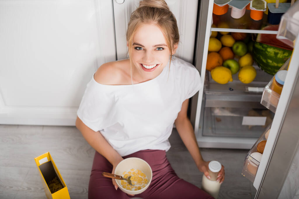 femme blonde assise sur le sol près du réfrigérateur ouvert avec bol de flocons de maïs et bouteille de lait - Photo, image