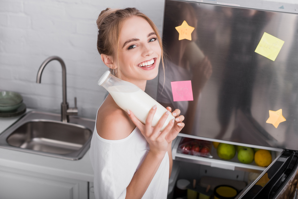 幸せなブロンド女性笑顔カメラで保持しながら牛乳のボトル近く冷蔵庫 - 写真・画像