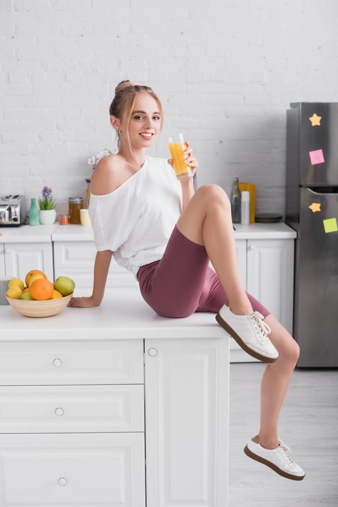 mulher loira em camiseta branca e shorts sentados na mesa na cozinha com copo de suco de laranja - Foto, Imagem