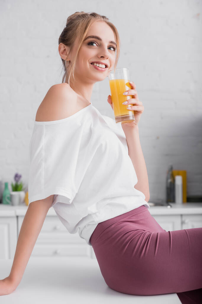 mosolygós szőke nő fehér pólóban kezében pohár narancslé, miközben ül a konyhaasztalon - Fotó, kép