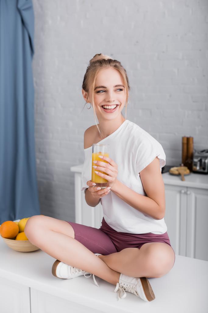 glückliche junge Frau im weißen T-Shirt, die mit überkreuzten Beinen auf dem Küchentisch sitzt und Orangensaft in der Hand hält - Foto, Bild