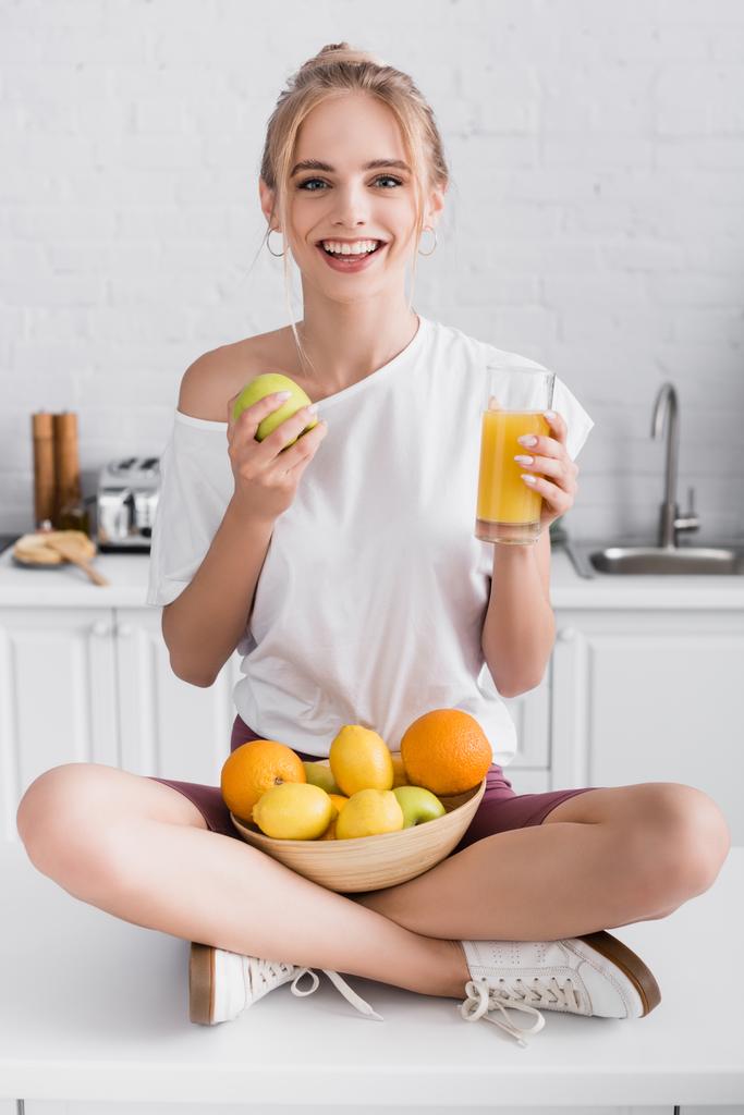 ευχαριστημένη νεαρή γυναίκα χαμογελώντας στην κάμερα, ενώ κάθεται στο τραπέζι της κουζίνας με φρούτα και ένα ποτήρι χυμό πορτοκαλιού - Φωτογραφία, εικόνα