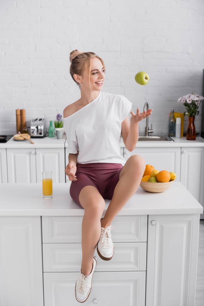 happy blonde woman juggling with apple while sitting on table in kitchen - Photo, Image