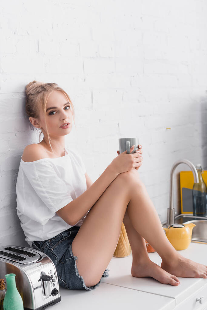 お茶を飲みながらキッチンテーブルに座りながらカメラを見ている官能的な素足の女性 - 写真・画像