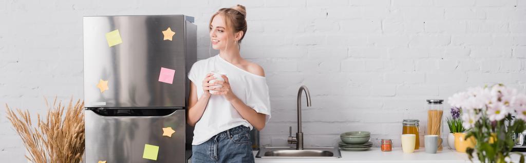 tiro panorâmico de jovem mulher de pé perto da geladeira na cozinha com xícara de chá e olhando para longe - Foto, Imagem