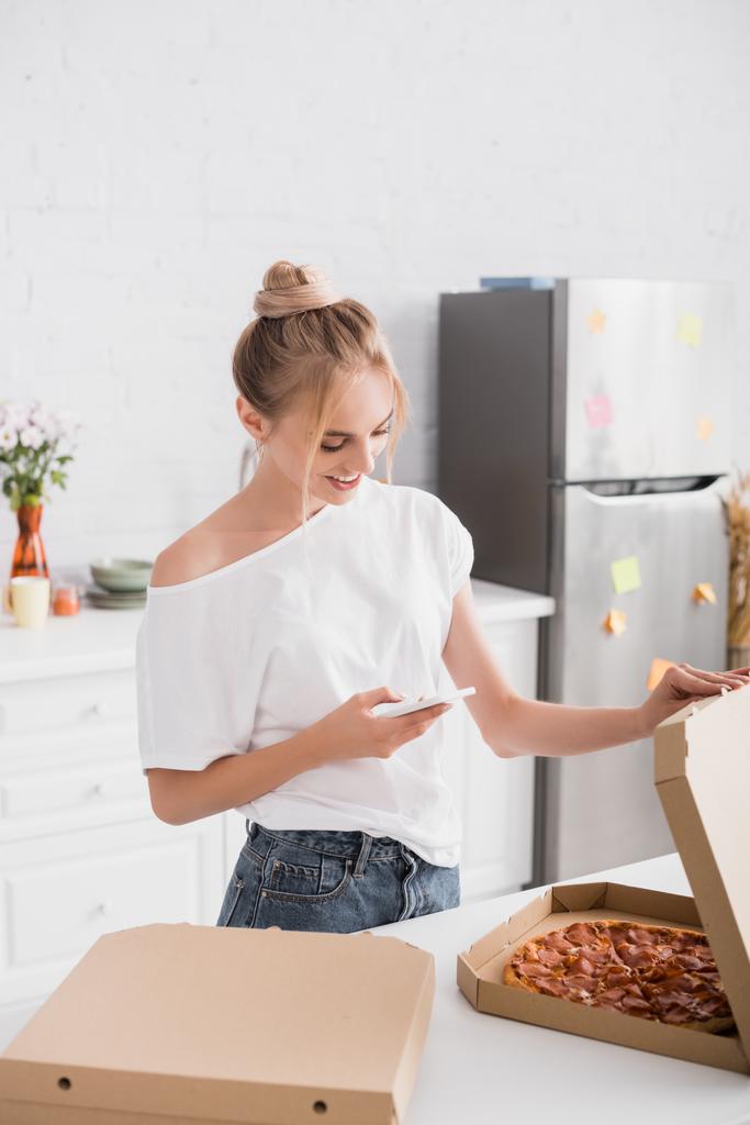 νεαρή γυναίκα σε λευκό t-shirt μηνυμάτων στο κινητό τηλέφωνο, ενώ το άνοιγμα κουτί με πίτσα στην κουζίνα - Φωτογραφία, εικόνα