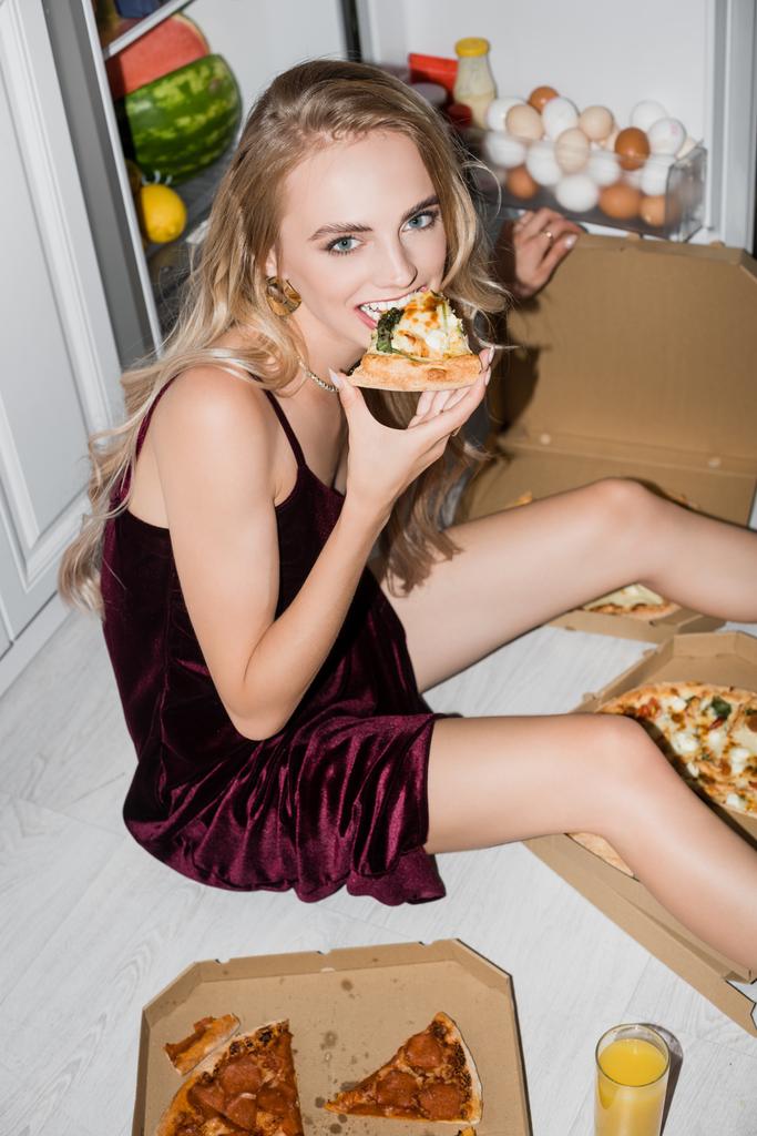 σέξι γυναίκα σε βελούδινο φόρεμα τρώει πίτσα, ενώ κάθεται στο πάτωμα κοντά άνοιξε ψυγείο - Φωτογραφία, εικόνα