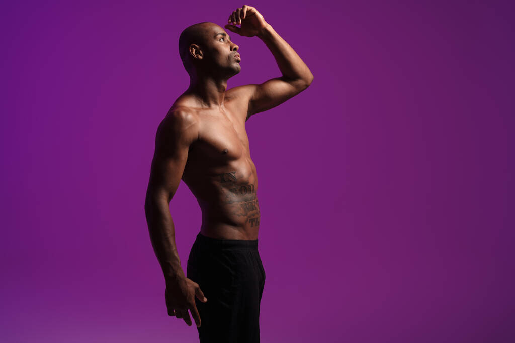 Imagen de atlético afroamericano semidesnudo con tatuaje posando en cámara aislada sobre pared púrpura - Foto, Imagen