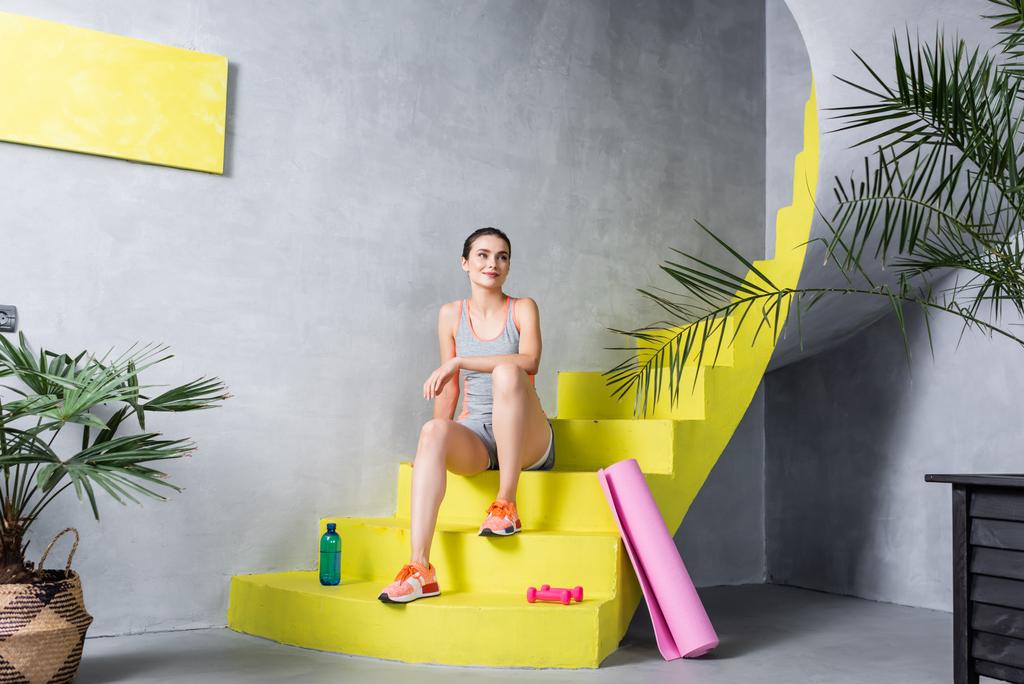 Женщина смотрит в сторону и сидит на лестнице возле спортивного инвентаря - Фото, изображение