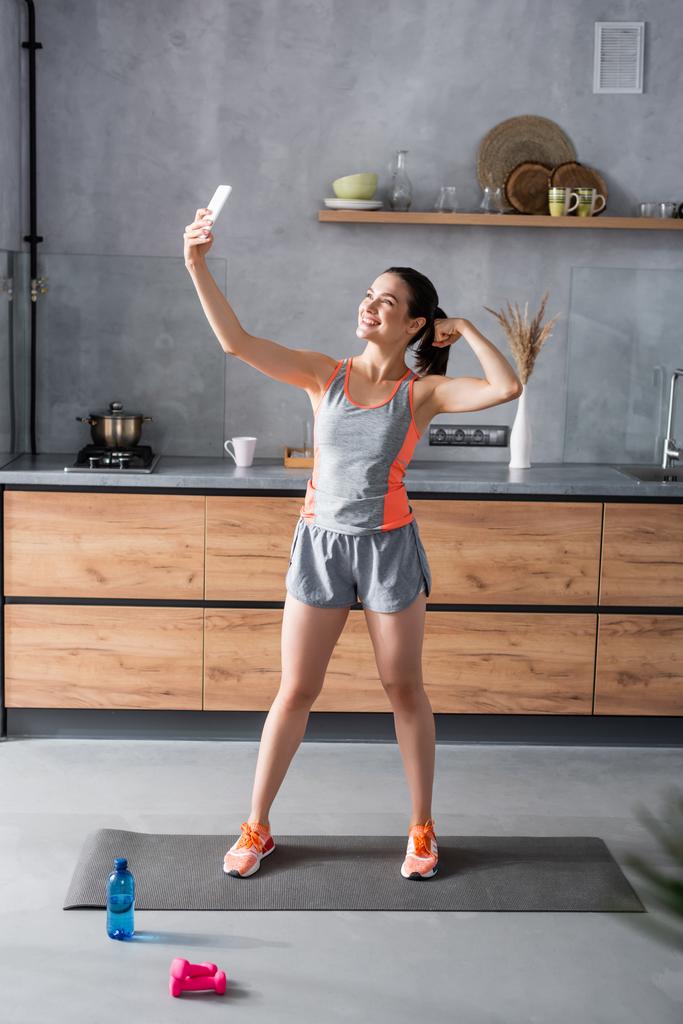Sporcu kadının evde su ve spor aletleri şişesinin yanındaki akıllı telefondan selfie çektiği seçici bir odak noktası.  - Fotoğraf, Görsel