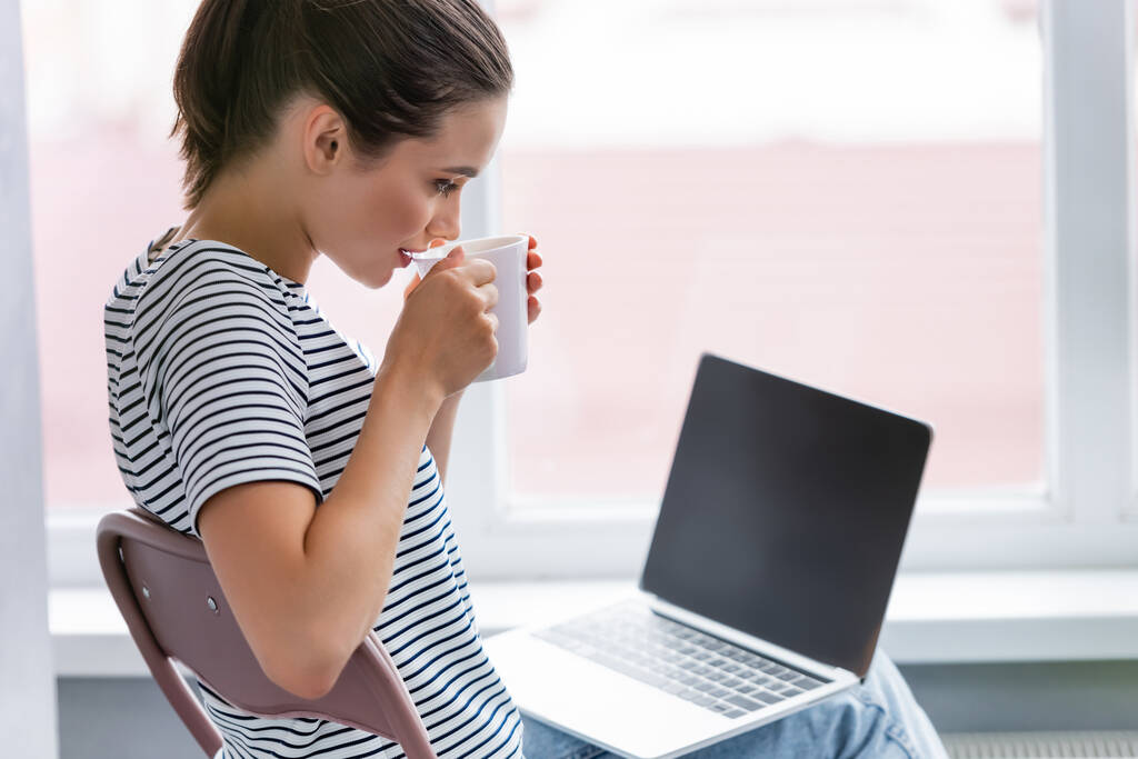 Válogatott fókusz a nő ivás tea és nézi laptop üres képernyőn otthon  - Fotó, kép
