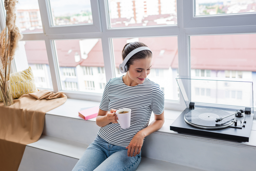 Mujer con taza escuchando música en auriculares cerca de reproductor de vinilo en el alféizar de la ventana  - Foto, Imagen