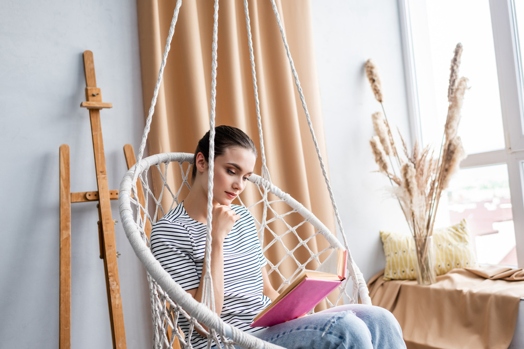 Jovem mulher lendo livro enquanto sentado em poltrona pendurada em casa  - Foto, Imagem